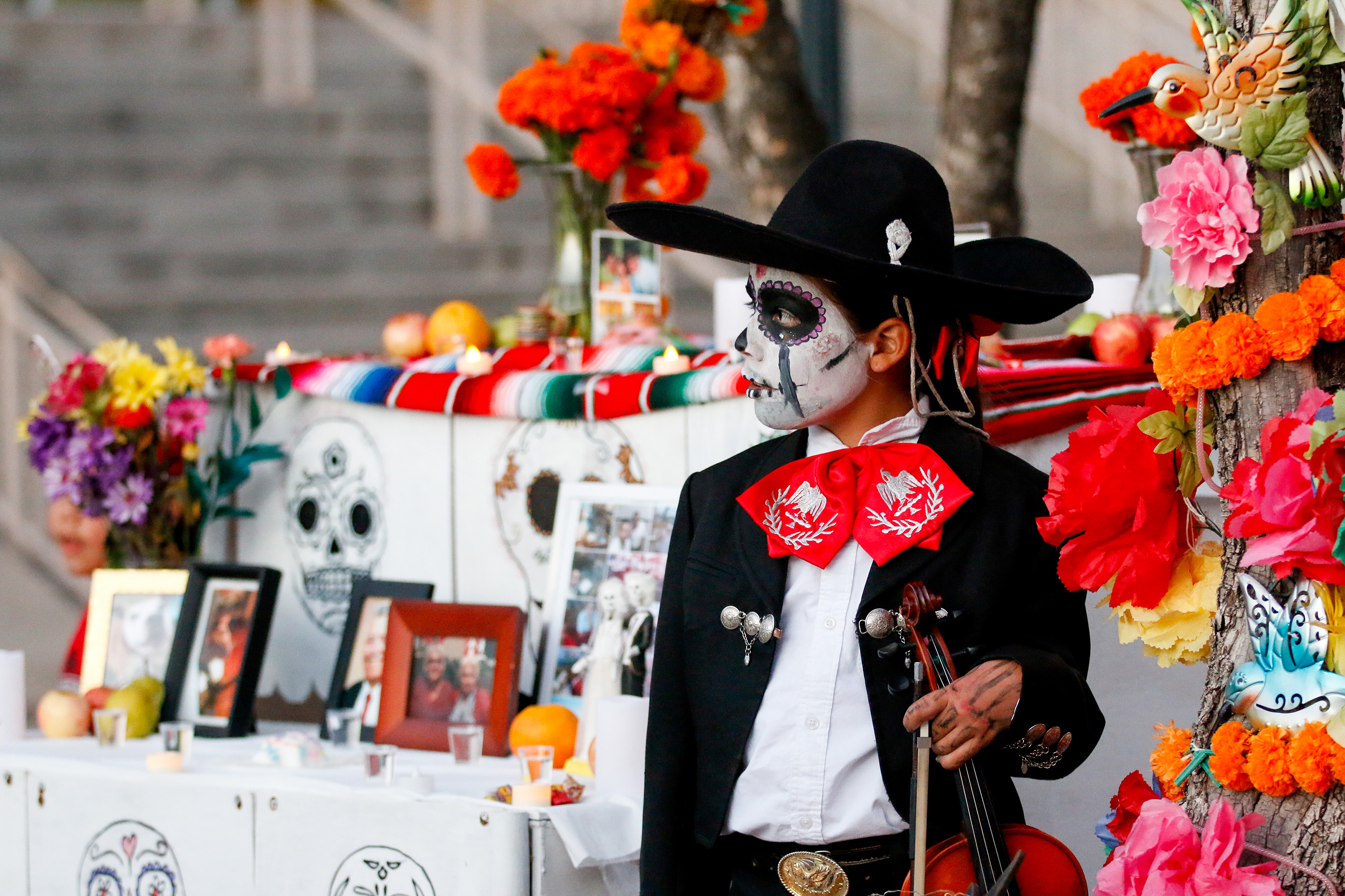 A boy wearing face paint and a mariachi costume holds his violin next to an ofrenda