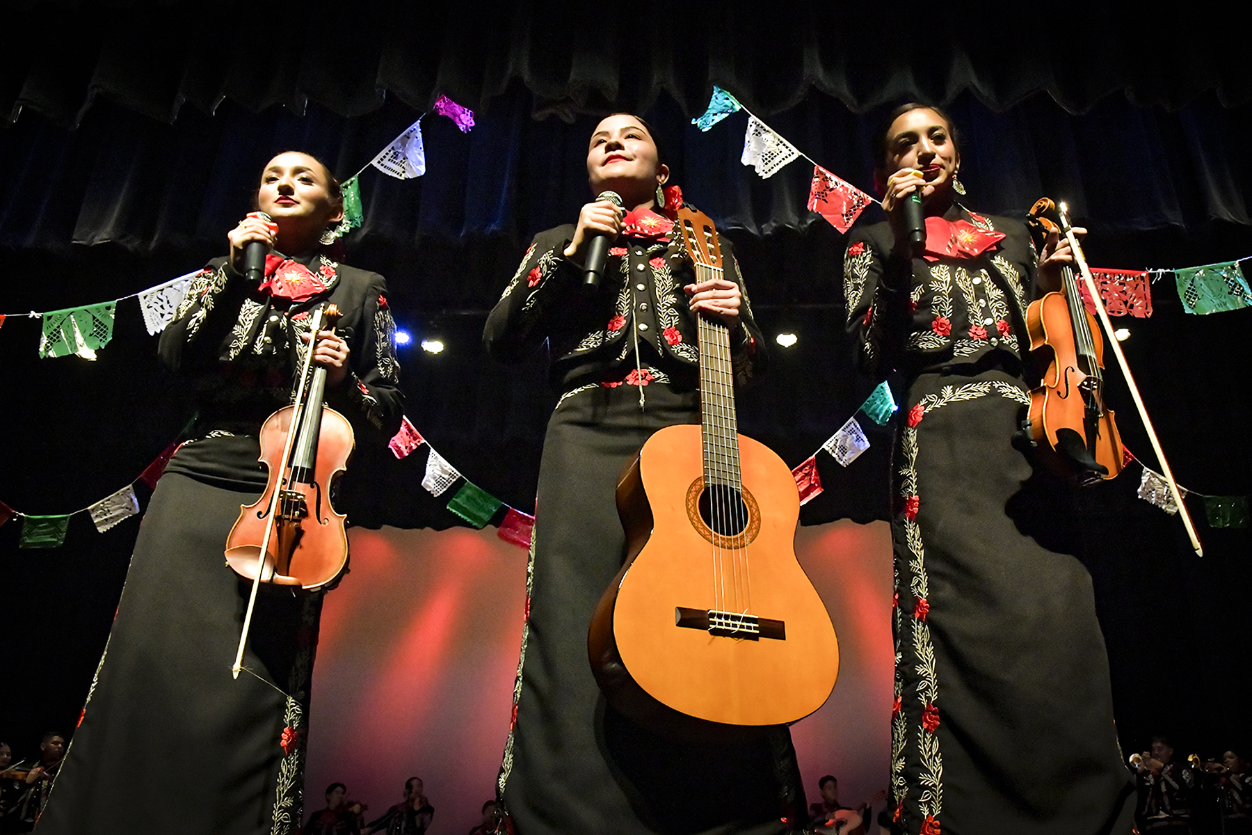 Three girls hold their instruments and microphones at the front of the stage