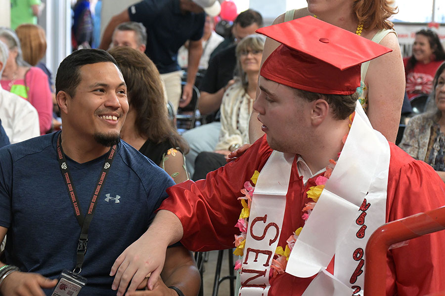 A teen boy in a red cap and gown talks to a man next to him