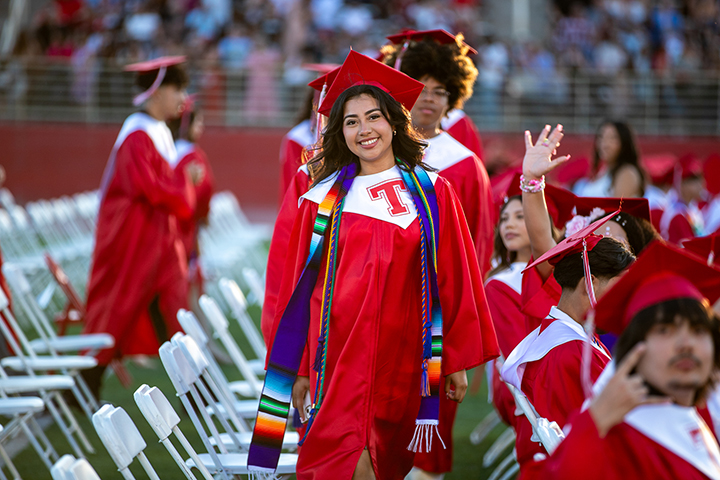 A teen girl in her red cap and gown smiles as she walks to her seat