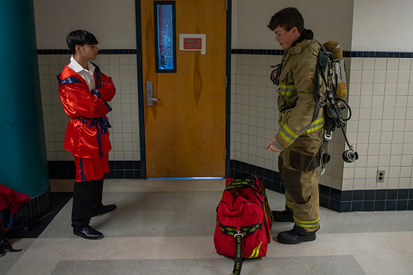 A student in firefighter gear talks to a student in a red robe and pants