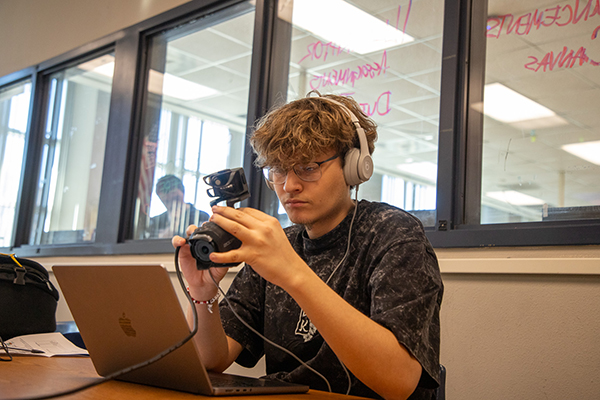 A teen boy holds up a camera as he sits in front of a computer