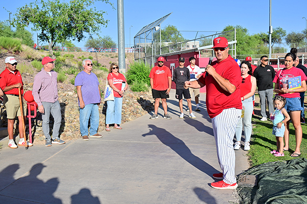 A man in a red and white baseball umpire uniform talks to the crowd outside the baseball field