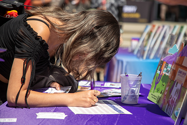 A teen girl leans over a table to sign a paper
