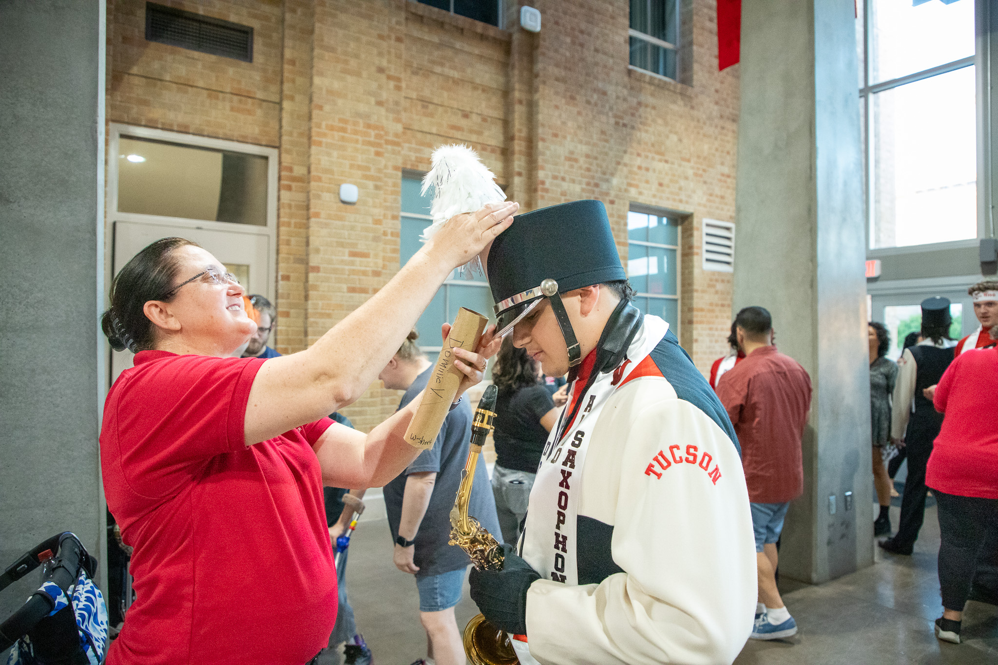 A woman in a red shirt adjusts a feather on a teen boy's marching band hat