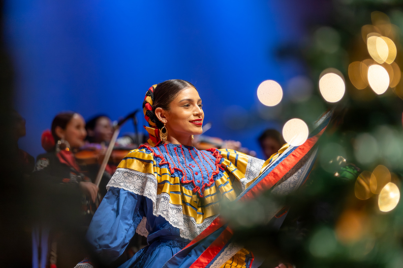 A girl in a colorful folklorico dress smiles as she dances on stage