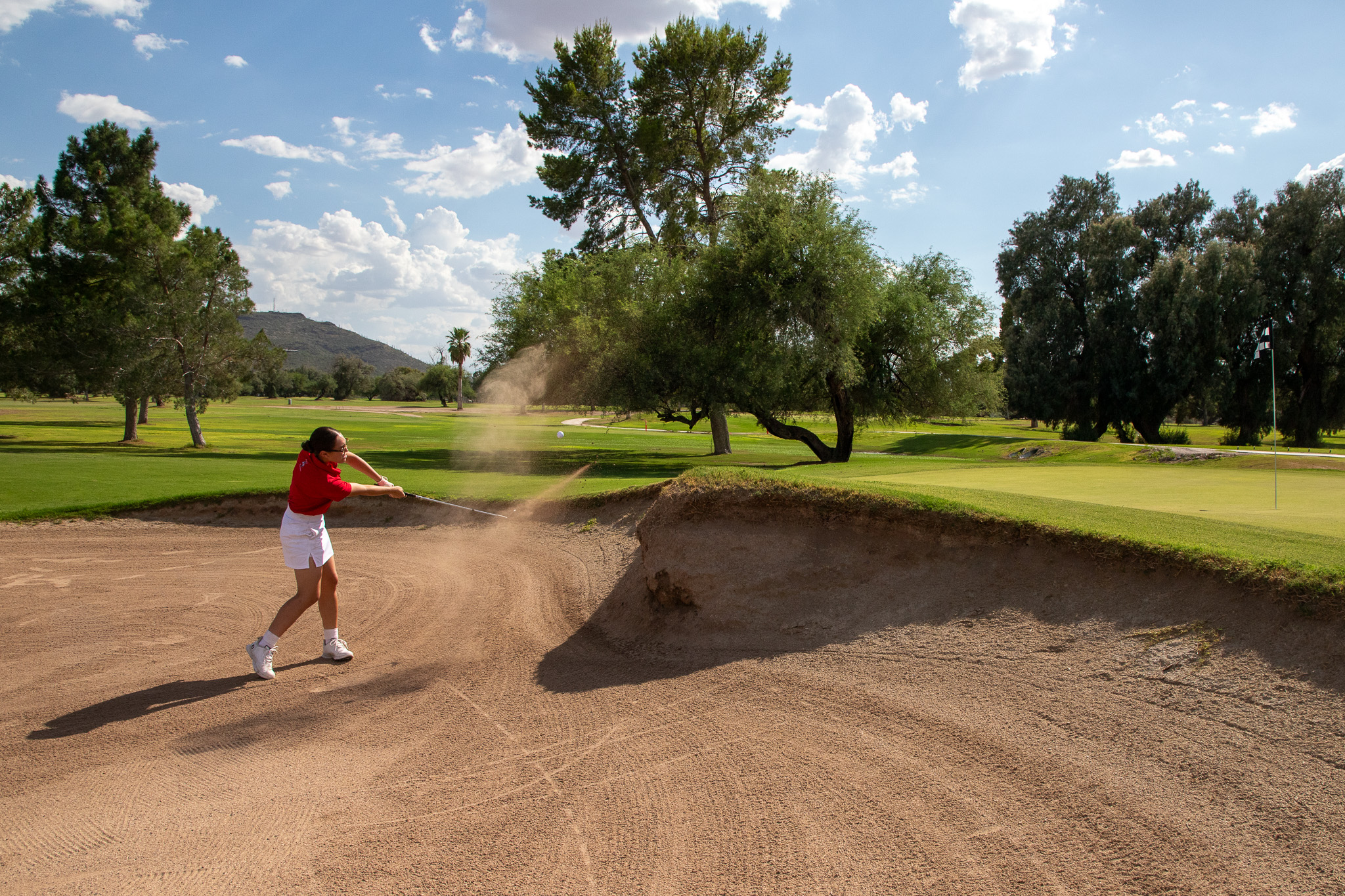 A teen girl swings her golf club to hit the ball out of the sand trap