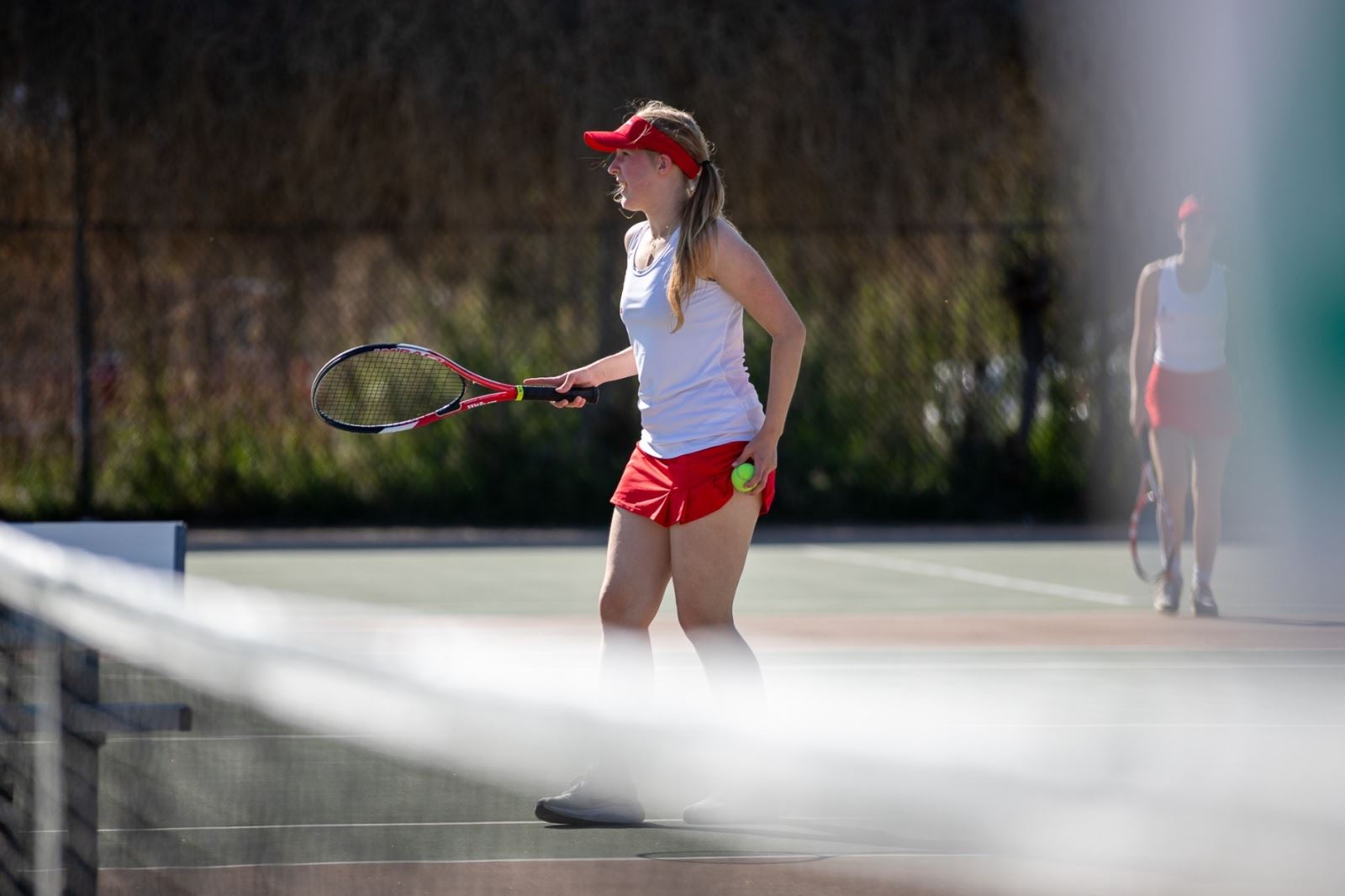 A THMS girls tennis player stands near the net holding a ball and racuqet.